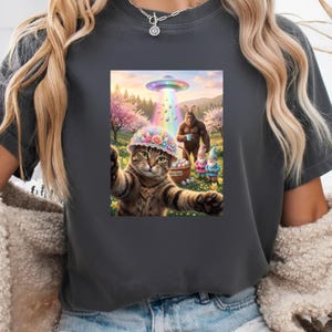 May include: Dark gray t-shirt featuring a whimsical graphic. The graphic depicts a cat wearing a floral hat taking a selfie with a UFO, Bigfoot, and Easter gnomes in a colorful spring landscape. A basket of Easter eggs is also visible.