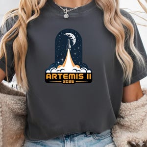 Puede incluir: Camiseta gris oscuro con un gráfico de un cohete lanzándose al espacio, con una luna y estrellas de fondo. El texto "ARTEMIS II 2026" se muestra en naranja y blanco.