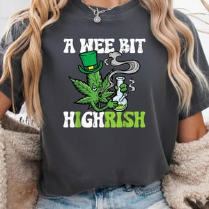 Könnte beinhalten: Dunkelgraues T-Shirt mit dem Text "A Wee Bit Highrish". Die Grafik zeigt ein grünes Cannabisblatt mit grünem Hut, das aus einer Bong raucht. Der Text "Highrish" ist in Weiß und Grün gehalten.