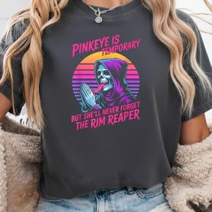 Może przedstawiać: Ciemnoszara koszulka z grafiką szkieletu w fioletowej szacie z kapturem, z wystawionym językiem, na tle retro zachodu słońca. Tekst brzmi: "Pinkeye is Temporary But She'll Never Forget The Rim Reaper."