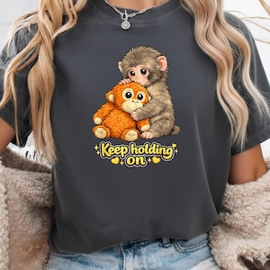 Puede incluir: Camiseta gris oscuro con una ilustración de dibujos animados de dos monos abrazándose. Un mono es marrón, el otro naranja. El texto "Keep holding on" está debajo de los monos, con corazones amarillos.