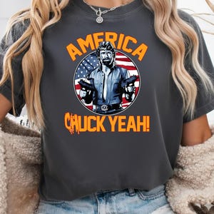 May include: Dark gray t-shirt featuring a graphic of a man holding two guns in front of an American flag. The text "AMERICA" and "CHUCK YEAH!" are in orange.