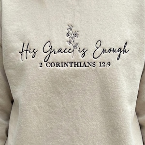 May include: Beige sweatshirt with embroidered text "His Grace is Enough" and "2 Corinthians 12:9". The design includes a small floral element above the text. The sweatshirt is a neutral color and has a crew neck.