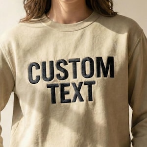 May include: A tan-colored sweatshirt with the words "CUSTOM TEXT" embroidered in black block letters across the chest. The sweatshirt has a crew neck and long sleeves. The fabric appears to be a soft, comfortable material.