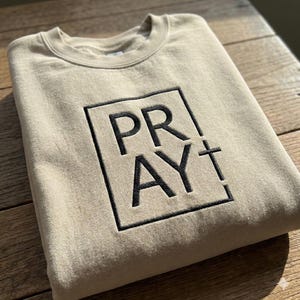 May include: A beige sweatshirt with the word "PRAY" embroidered in black within a square, with a cross symbol. The sweatshirt is made of a soft material and is suitable for casual wear.