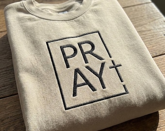 May include: A beige sweatshirt with the word "PRAY" embroidered in black within a square, with a cross symbol. The sweatshirt is made of a soft material and is suitable for casual wear.