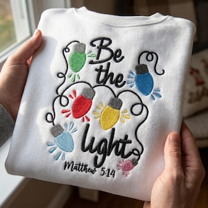 以下が含まれることがあります： Be the lightおよびMatthew 5:14と刺繍された白いスウェットシャツ。デザインには、黒い糸で刺繍されたライトの紐が含まれており、緑、青、赤、黄色、ピンクのカラフルな電球が並び、それぞれに小さな放射状の線が施されています。