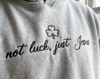 May include: A gray hoodie with a black embroidered shamrock and the phrase "not luck, just Jesus." The hoodie has a drawstring hood and a kangaroo pocket. The text is in a cursive font.