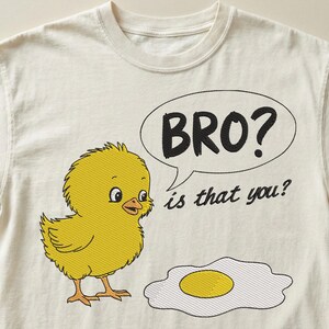 May include: Cream-colored t-shirt featuring an embroidered yellow chick with the text "BRO? is that you?" in a speech bubble. An embroidered fried egg is also present. The design is whimsical and playful.