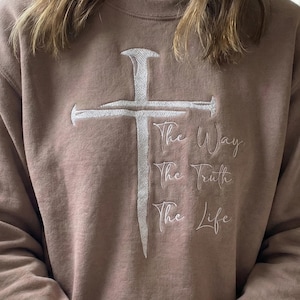 Puede incluir: Sudadera color canela con un diseño de cruz bordada en blanco. Debajo de la cruz, las palabras "The Way, The Truth, The Life" están bordadas en letra cursiva. La sudadera tiene cuello redondo y mangas largas.