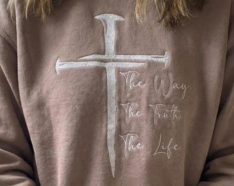 May include: A tan sweatshirt featuring a white embroidered cross design. Below the cross, the words "The Way, The Truth, The Life" are embroidered in a script font. The sweatshirt has a crew neck and long sleeves.