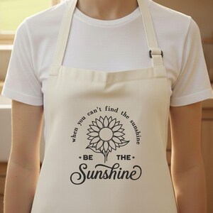 May include: Cream apron featuring a black sunflower design and the words "When you can't find the sunshine, be the sunshine." It has adjustable straps and a front pocket. The apron is made of a durable fabric.