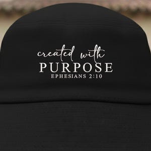 May include: Black baseball cap with the words "created with PURPOSE" in white script and block letters. The text "Ephesians 2:10" is below the word "PURPOSE". The cap is made of a textured fabric.