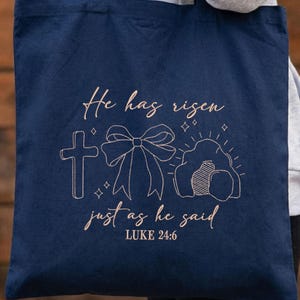 May include: Navy blue tote bag with a light brown embroidered design. The design includes a cross, a bow, and an empty tomb, with the text "He has risen just as he said" and "LUKE 24:6".