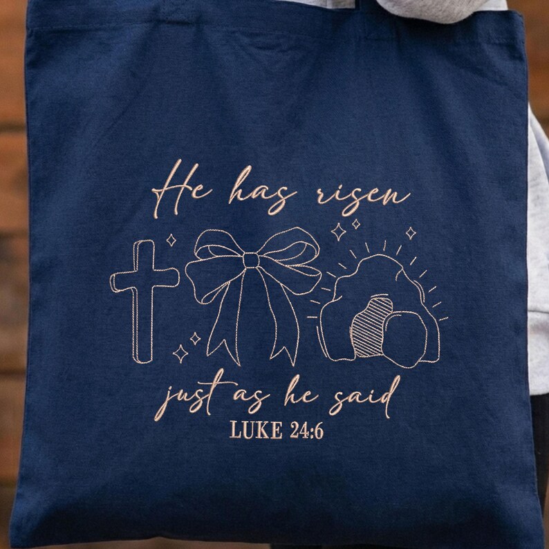 May include: Navy blue tote bag with a light brown embroidered design. The design includes a cross, a bow, and an empty tomb, with the text "He has risen just as he said" and "LUKE 24:6".