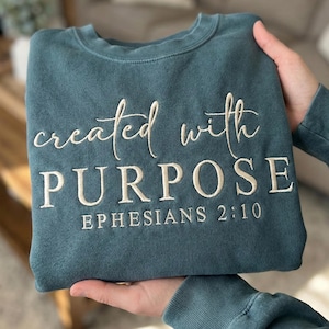 Peut inclure: Sweat-shirt bleu sarcelle avec l'inscription brodée en blanc "created with PURPOSE" et "EPHESIANS 2:10". Le sweat-shirt à col rond est fait d'une matière douce.
