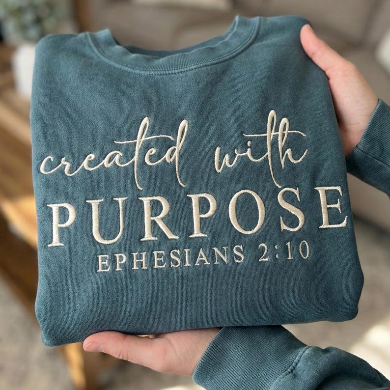 May include: Teal-colored sweatshirt with the embroidered text "created with PURPOSE" and "EPHESIANS 2:10" in white. The sweatshirt is made of a soft material and has a crew neck.