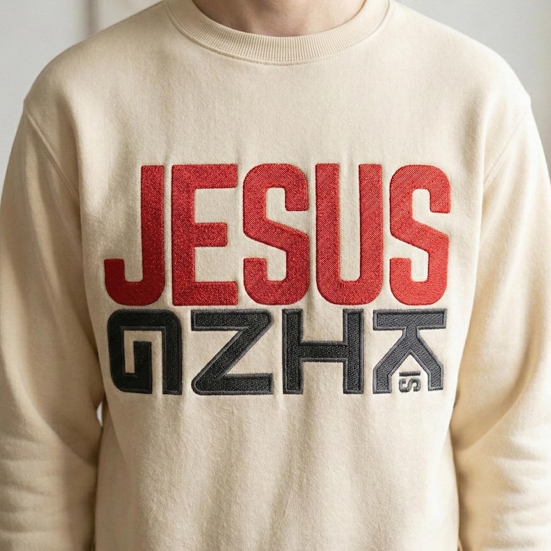 May include: Cream-colored sweatshirt with the word "JESUS" in red, embroidered lettering. Below, the words "GZHK IS" are embroidered in black. The sweatshirt has a crew neck and a relaxed fit.