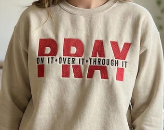 May include: A beige sweatshirt with the word "PRAY" in large, red, embroidered letters across the chest. Below the word "PRAY" are the words "ON IT • OVER IT • THROUGH IT" in smaller, red letters.