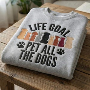 May include: A grey sweatshirt featuring the embroidered phrase "LIFE GOAL PET ALL THE DOGS." The design incorporates paw prints and dog silhouettes in brown, grey, and black, perfect for dog lovers.