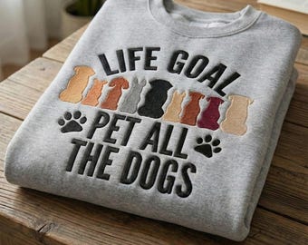 May include: A gray sweatshirt with the embroidered text "LIFE GOAL PET ALL THE DOGS." The design includes paw prints and silhouettes of various dog breeds in shades of brown, gray, and black.