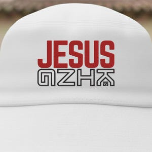 May include: White baseball cap with the word "JESUS" in red block letters above a black and white graphic. The cap is made of a textured fabric.