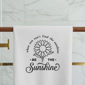 May include: White towel featuring a sunflower design and the words "When you can't find the sunshine, be the sunshine." The towel is displayed on a wooden rail in a bathroom.