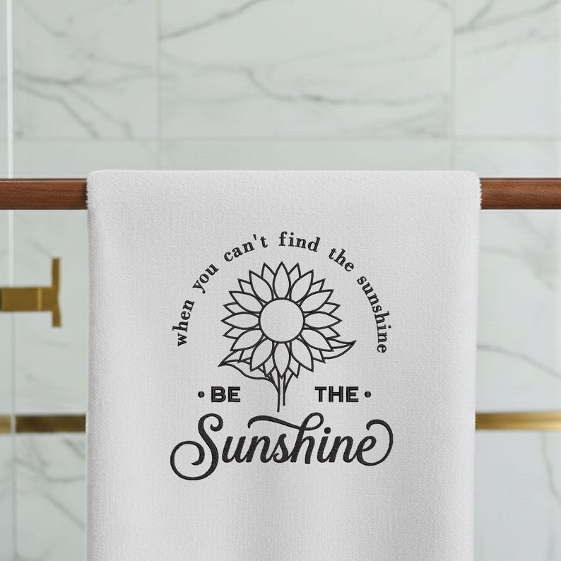 May include: White towel featuring a sunflower design and the words "When you can't find the sunshine, be the sunshine." The towel is displayed on a wooden rail in a bathroom.