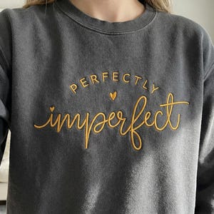 Op de afbeelding: Donkergrijze sweatshirt met de woorden "perfectly imperfect" geborduurd in goudkleurig garen. De tekst is in een cursief lettertype met een klein hartsymbool. De sweatshirt heeft een ronde hals.