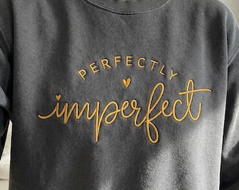 May include: Dark gray sweatshirt with the words "perfectly imperfect" embroidered in gold thread. The text is in a cursive font with a small heart symbol above the word "imperfect". The sweatshirt has a crew neck.