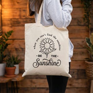 May include: A cream-coloured tote bag featuring a sunflower design and the words "When you can't find the sunshine, be the sunshine." The bag has two handles and is made of a durable fabric. The design is in black, with a simple, elegant font.