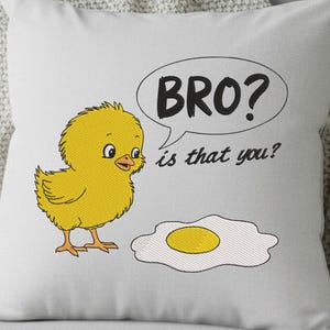 May include: A white decorative pillow with an embroidered design. A yellow chick looks at a fried egg. A speech bubble says "BRO? is that you?" in black text. The chick and egg are yellow, orange, and white.