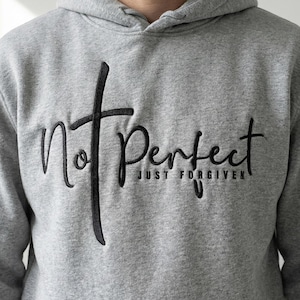 Könnte beinhalten: Ein grauer Kapuzenpullover mit den gestickten Wörtern "Not Perfect" und "Just Forgiven" in schwarzer Schrift. Das Wort "Not" ist mit einem Kreuz versehen. Der Kapuzenpullover hat eine Kapuze und ein weiches, bequemes Aussehen.