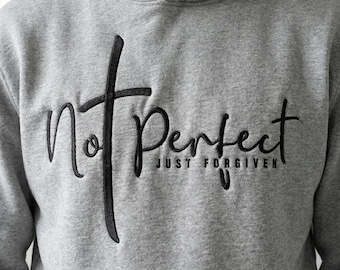 May include: A gray hoodie with the embroidered text "Not Perfect" and "Just Forgiven" in black script. The word "Not" is stylized with a cross. The hoodie has a hood and a soft, comfortable appearance.