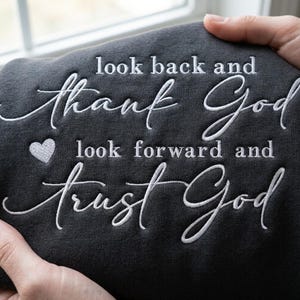 May include: A dark charcoal gray sweatshirt featuring white embroidered text that reads look back and thank God, look forward and trust God, with a small embroidered heart icon positioned between the two lines of text on the left side.