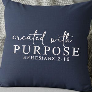 May include: Navy blue square pillow with the words "created with PURPOSE" in white script and block letters. The text "EPHESIANS 2:10" is below. The pillow is resting on a gray surface.