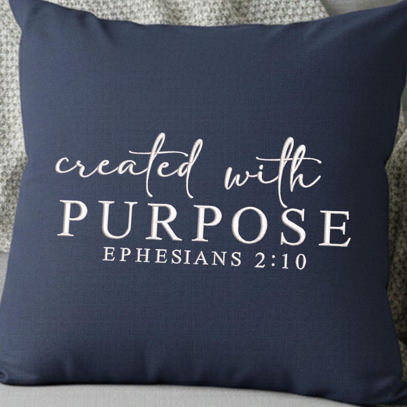 May include: Navy blue square pillow with the words "created with PURPOSE" in white script and block letters. The text "EPHESIANS 2:10" is below. The pillow is resting on a gray surface.