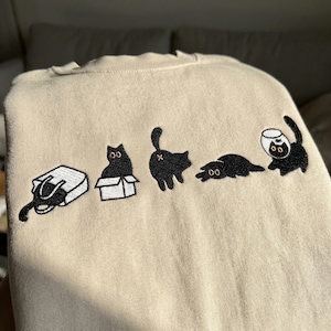 Pode incluir: Camisola bege com desenhos de gatos pretos bordados. Os gatos são mostrados em diferentes poses, incluindo dentro de uma caixa, deitados e com um aquário. A camisola tem gola redonda e mangas compridas.