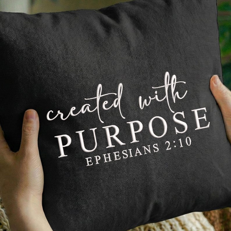 May include: Black square pillow with white text that reads "created with PURPOSE EPHESIANS 2:10". The pillow is held by a person. The text is in a mix of cursive and block letters.