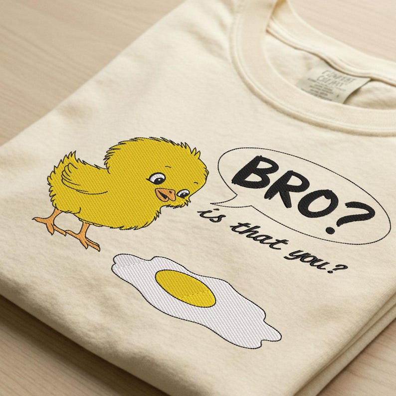 May include: Cream-colored t-shirt featuring a yellow chick looking at a fried egg. A speech bubble says "BRO? is that you?" in black text. The shirt is folded, showing the collar and a tag.