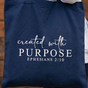 May include: Navy blue tote bag with the words "created with PURPOSE" and "EPHESIANS 2:10" in white script and block letters. The bag is made of a soft fabric and is suitable for carrying books, groceries, or other items.