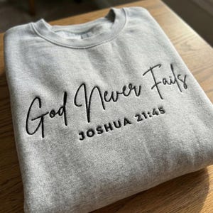 May include: A gray sweatshirt with the embroidered text "God Never Fails" and "Joshua 21:45" in black script. The sweatshirt is folded on a wooden surface. The crew neck sweatshirt is a casual style.