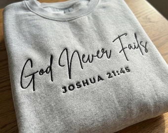 May include: A gray sweatshirt with the embroidered text "God Never Fails" and "Joshua 21:45" in black script. The sweatshirt is folded on a wooden surface. The crew neck sweatshirt is a casual style.