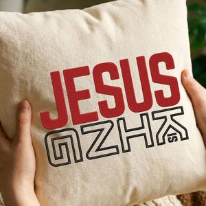 May include: Cream-colored square pillow with the word "JESUS" in red, bold letters. Below, the letters "GZHK" are outlined in black. The pillow is held by two hands.