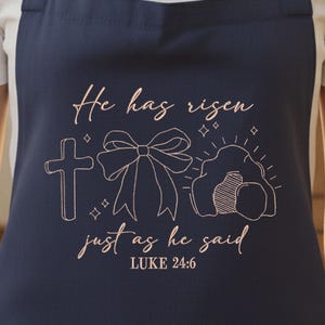 May include: Navy blue apron with a gold-colored embroidered design. The design includes a cross, a bow, and an empty tomb, with the words "He has risen just as he said" and the Bible verse Luke 24:6.