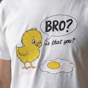 May include: White t-shirt with an embroidered design featuring a yellow chick, a fried egg, and a speech bubble with the text "BRO? is that you?". The chick has blue eyes and orange feet.