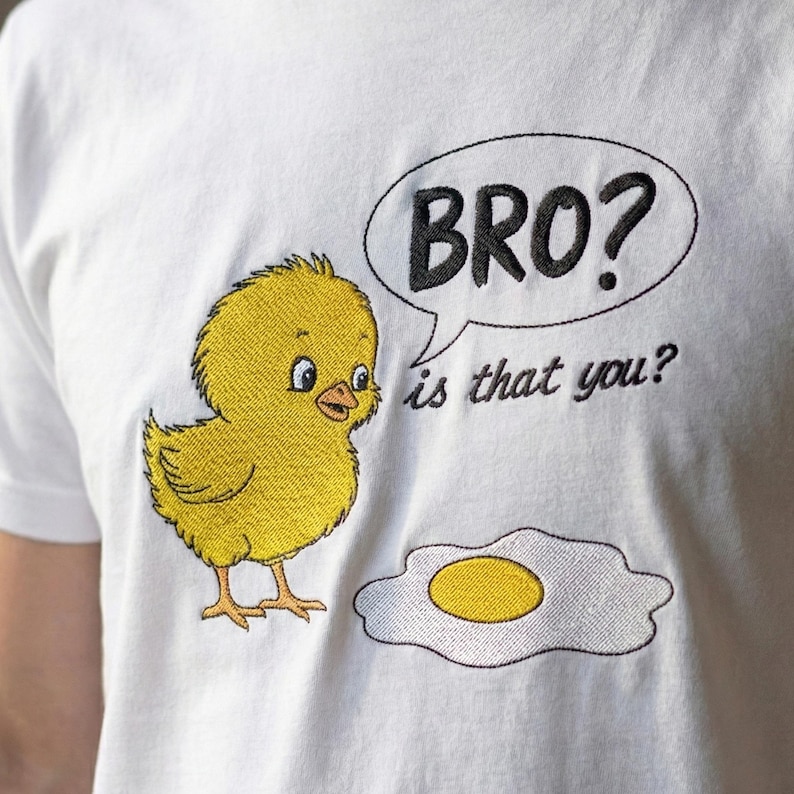May include: White t-shirt with an embroidered design featuring a yellow chick, a fried egg, and a speech bubble with the text "BRO? is that you?". The chick has blue eyes and orange feet.