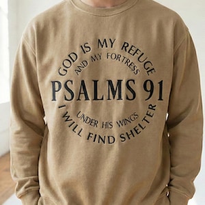 May include: A tan-colored sweatshirt with the text "GOD IS MY REFUGE AND MY FORTRESS PSALMS 91 I WILL FIND SHELTER UNDER HIS WINGS" printed in black. The text is arranged in a circular design.