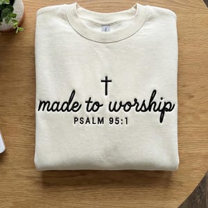 May include: Cream-colored sweatshirt with the embroidered text "made to worship" and a cross symbol. Below the text is "PSALM 95:1" in black thread. The sweatshirt is folded on a wooden surface.