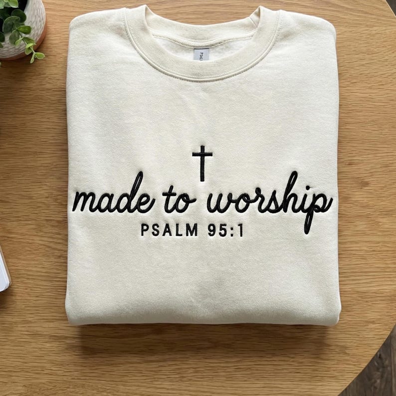 May include: Cream-colored sweatshirt with the embroidered text "made to worship" and a cross symbol. Below the text is "PSALM 95:1" in black thread. The sweatshirt is folded on a wooden surface.